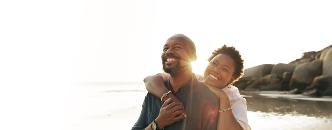 A smiling man and woman, walking along a sandy beach near the waterline, with the woman slightly behind the man, her arms around his shoulder; sun shining, small waves and open ocean in the background.
